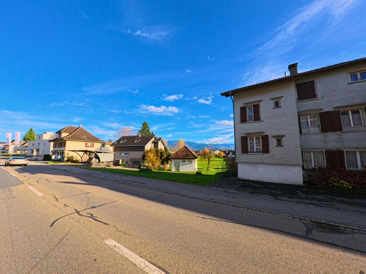 Mehrere Wohnhaeuser stehen in Marbach (9437, Kanton St. Gallen) entlang einer ruhigen Strasse, waehrend im Hintergrund gruene Wiesen und Berge sichtbar sind.