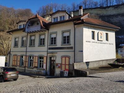Das Musée de la Marionnette in Fribourg (1701, Kanton Freiburg) befindet sich in einem hellen Gebäude mit roten Fensterläden an einer gepflasterten Strasse.