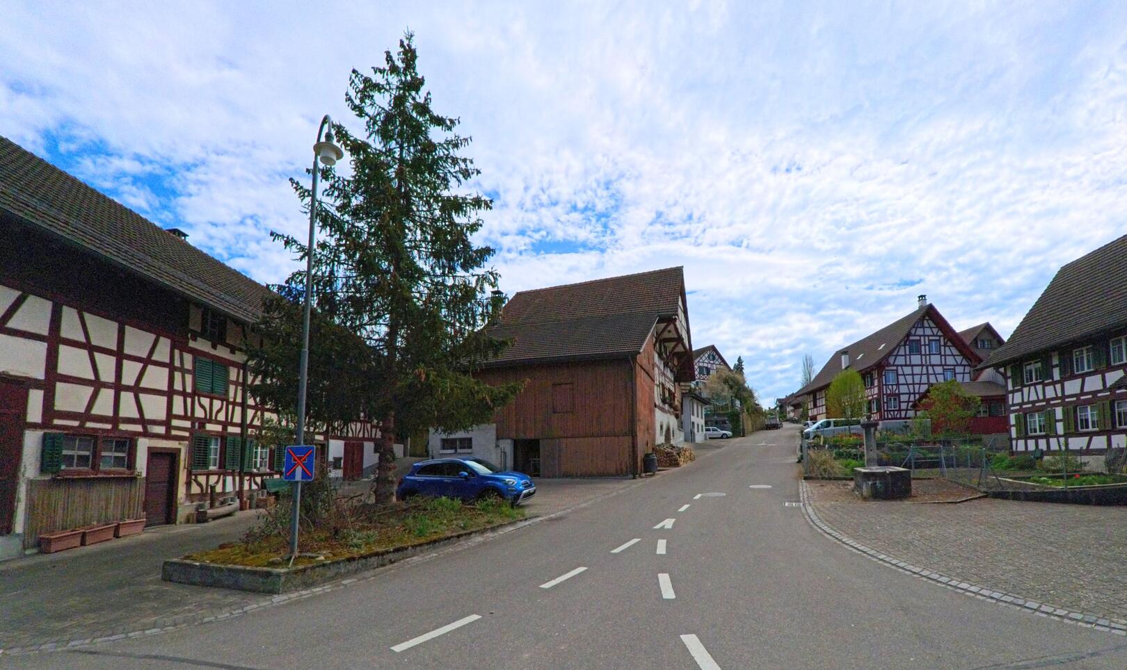 In Marthalen (8460, Kanton Zürich) führt eine ruhige Strasse an traditionellen Fachwerkhäusern entlang, während der Blick auf einen sanft ansteigenden Hügel fällt.