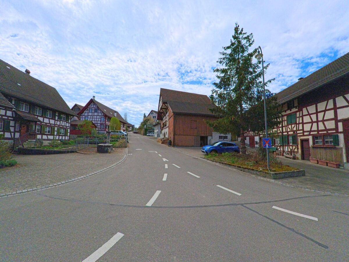 In Marthalen im Kanton Zürich mit der PLZ 8460 verläuft eine Hauptstrasse durch das Dorfzentrum, gesäumt von traditionellen Fachwerkhäusern und einem kleinen Baumbeet mit Strassenlaterne.
