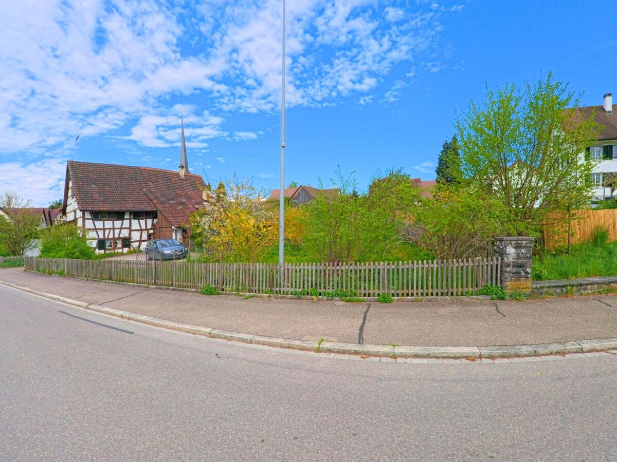 In Marthalen im Kanton Zürich mit der PLZ 8460 steht ein schmuckes Fachwerkhaus mit spitzem Dach und Kirchturm hinter einem blühenden Garten und Holzzaun an einer Kurve.