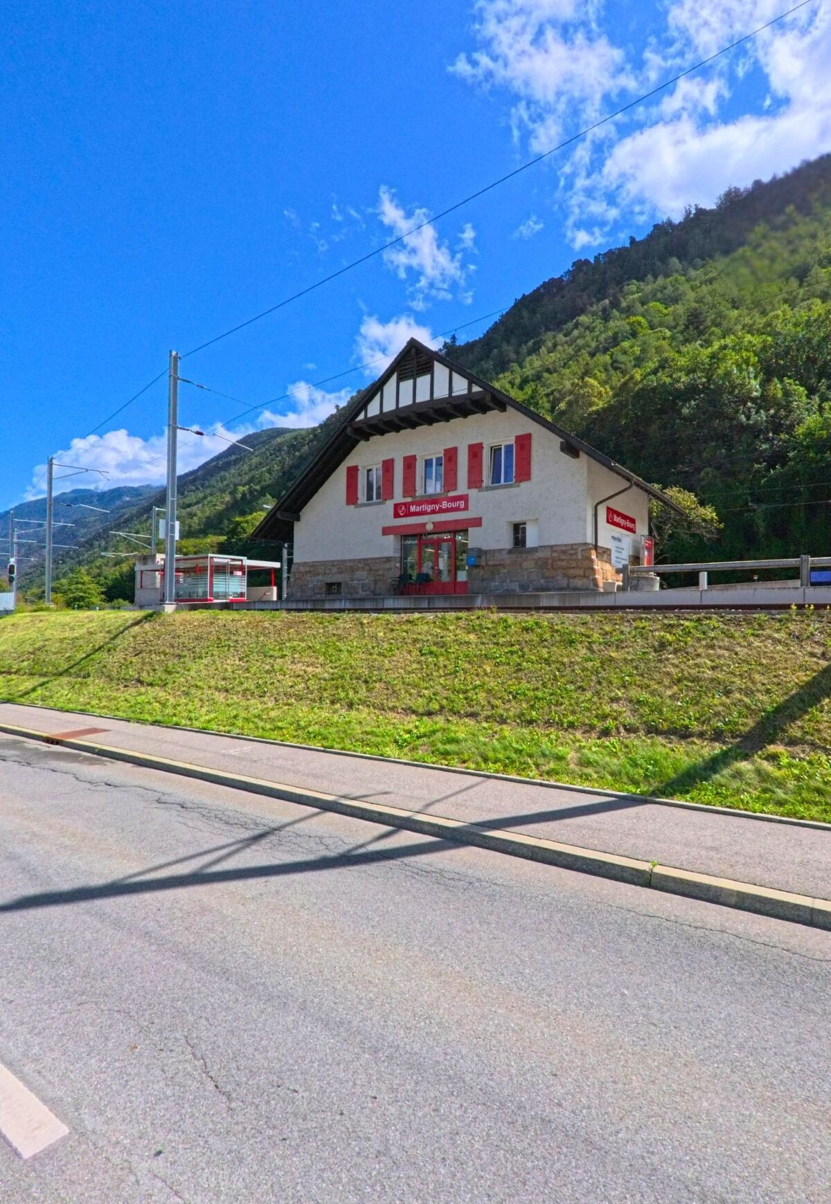 Das Bahnhofsgebäude Martigny-Bourg in Martigny (1920, Kanton Wallis) steht vor bewaldeten Hügeln und zeigt rote Fensterläden sowie ein markantes Satteldach.