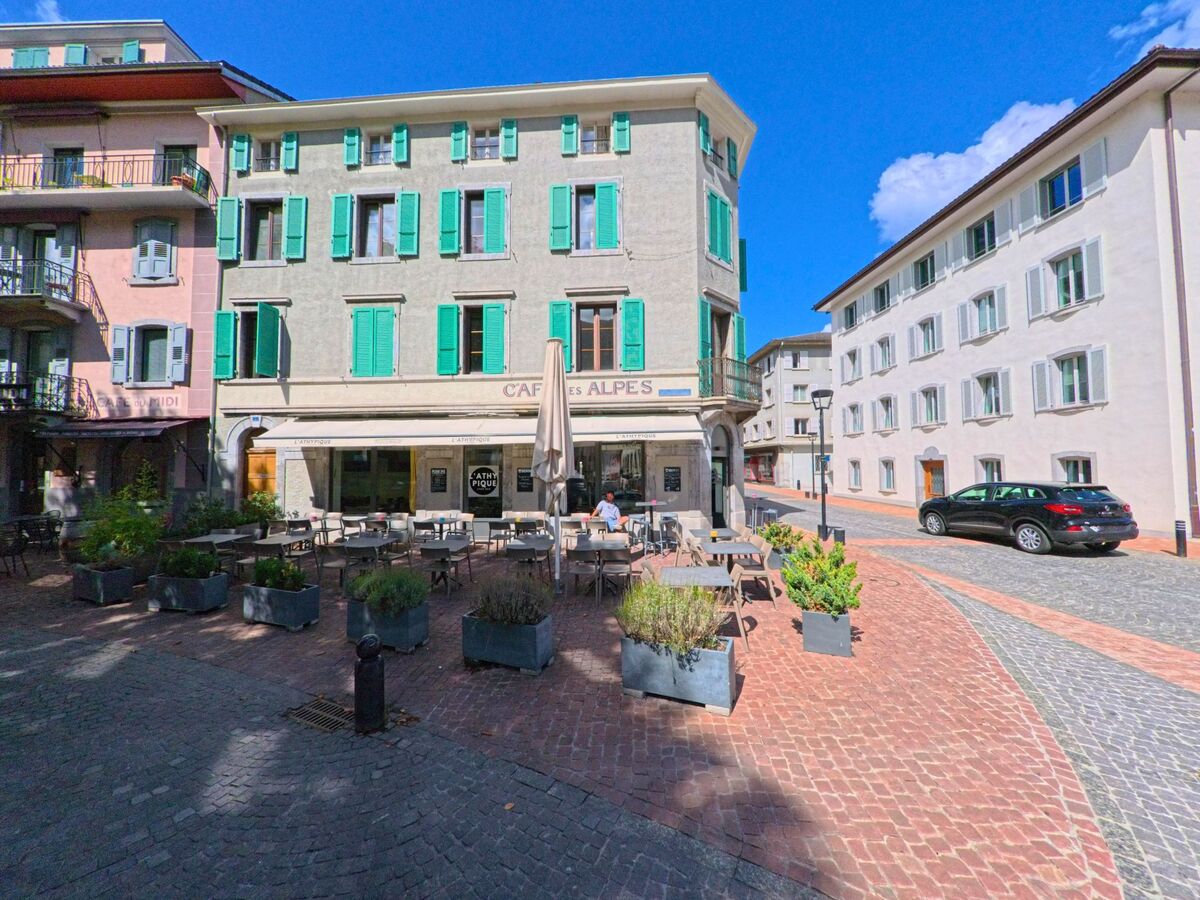 Vor dem Gebäude des Café des Alpes in Martigny (1920, Kanton Wallis) stehen zahlreiche Tische und Stühle auf einem gepflasterten Platz, eingerahmt von Wohnhäusern.