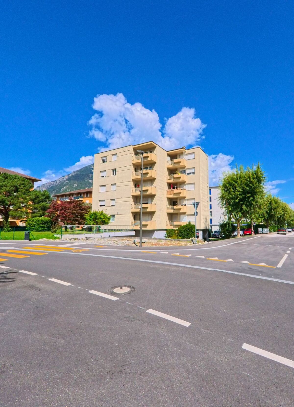 Ein helles Mehrfamilienhaus mit Balkonen steht an einer Kreuzung in Martigny (1920, Kanton Wallis), im Hintergrund sind Berge und weitere Gebäude zu sehen.