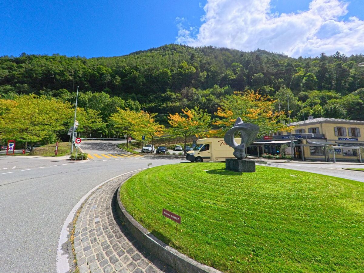 Auf einem Kreisverkehr in Martigny (1921, Kanton Wallis) steht eine moderne Steinskulptur, umgeben von Bäumen mit beginnender Herbstfärbung vor dem bewaldeten Hügel.