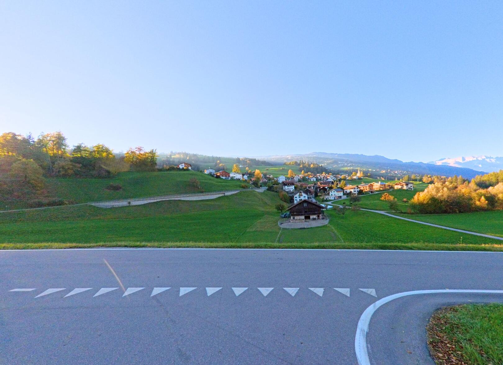 Ein weiter Blick über grüne Wiesen und verstreute Häuser zeigt das Dorf Masein (7425, Kanton Graubünden) mit einer geschwungenen Strasse im Vordergrund und Hügeln am Horizont.
