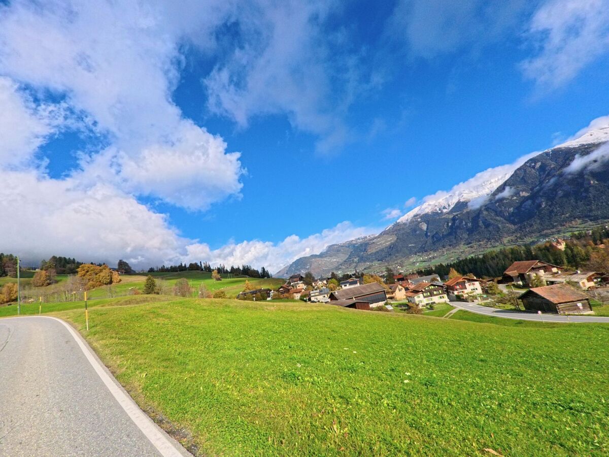 Von einer schmalen Strasse aus öffnet sich in Masein (7425, Kanton Graubünden) der Blick über weite Wiesen auf das Dorf und die markanten Berghänge unter einem wolkenreichen Himmel.