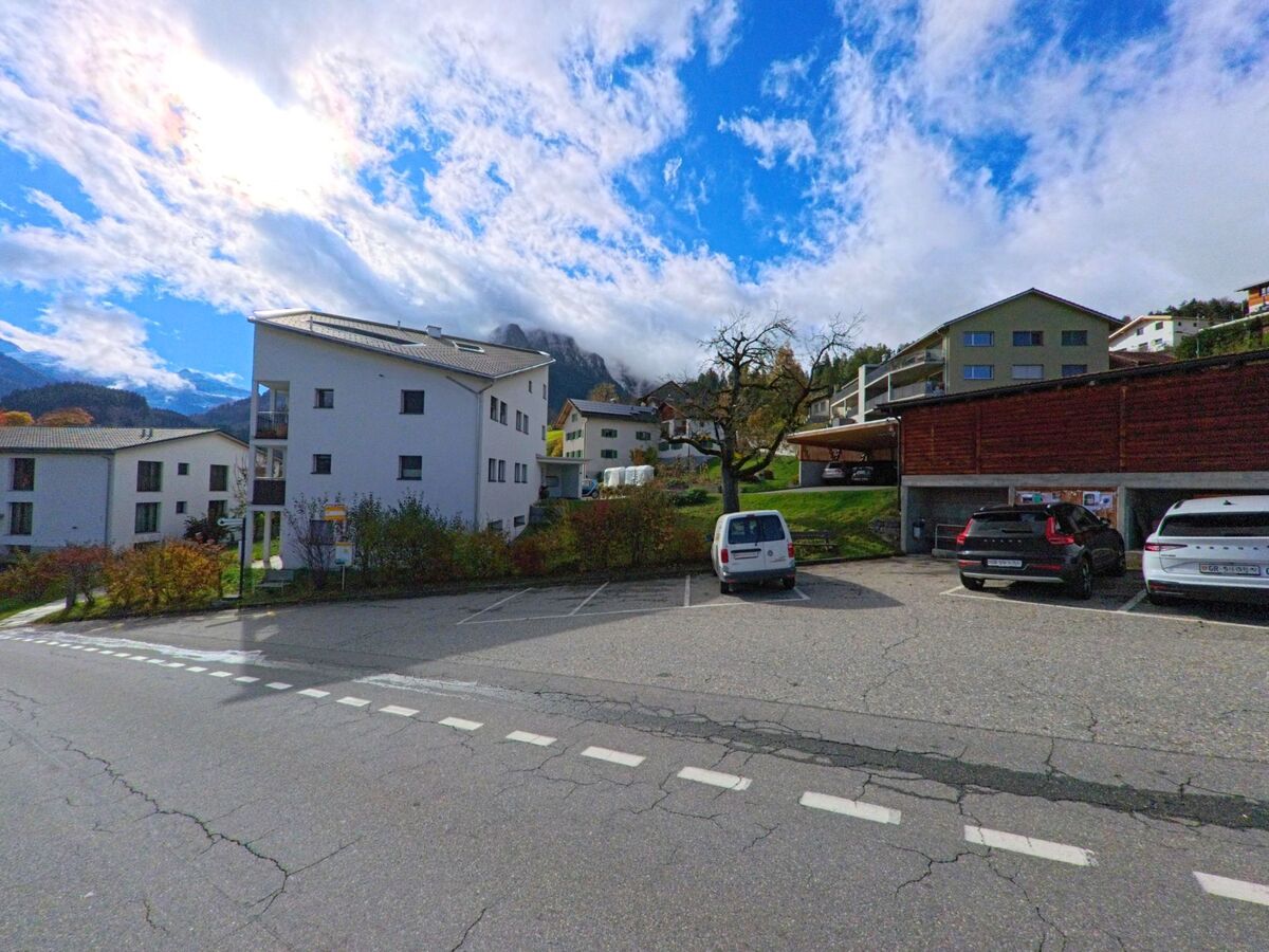 Vor modernen Wohnbauten und parkierenden Fahrzeugen zeigt sich in Masein (7425, Kanton Graubünden) eine ruhige Dorfstrasse mit Blick auf bewaldete Hänge und tief hängende Wolken.