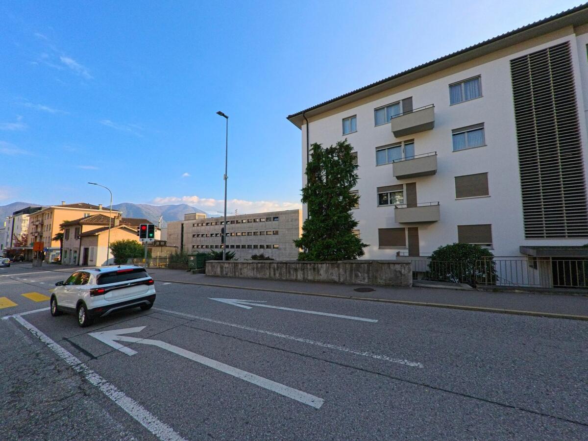 An einer Ampelkreuzung in Massagno (6900, Kanton Tessin) verläuft eine mehrspurige Strasse entlang von Wohnhäusern mit Blick auf die umliegenden Hügel.