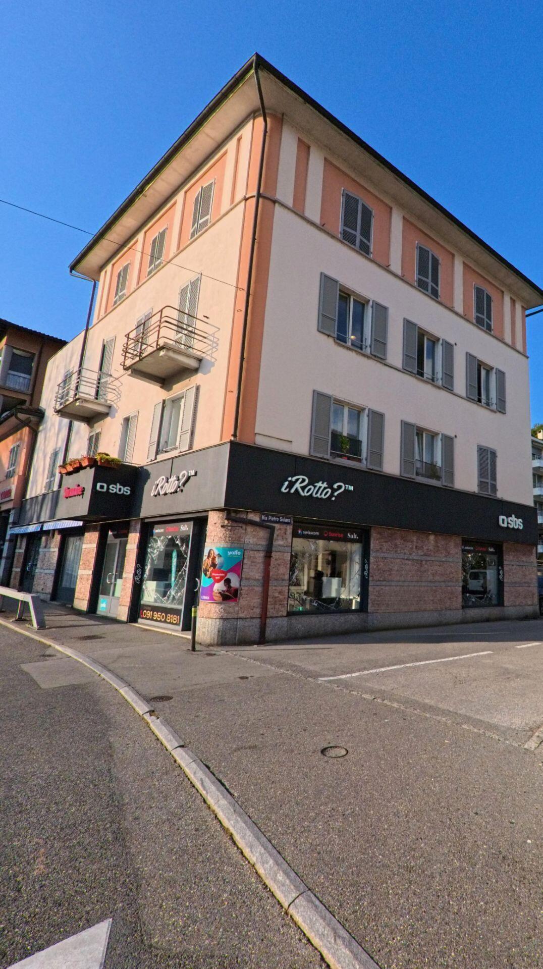 An einer Strassenecke in Massagno (6900, Kanton Tessin) steht ein mehrstöckiges Wohn- und Geschäftshaus mit Ladenlokalen im Erdgeschoss und Balkonen darüber.