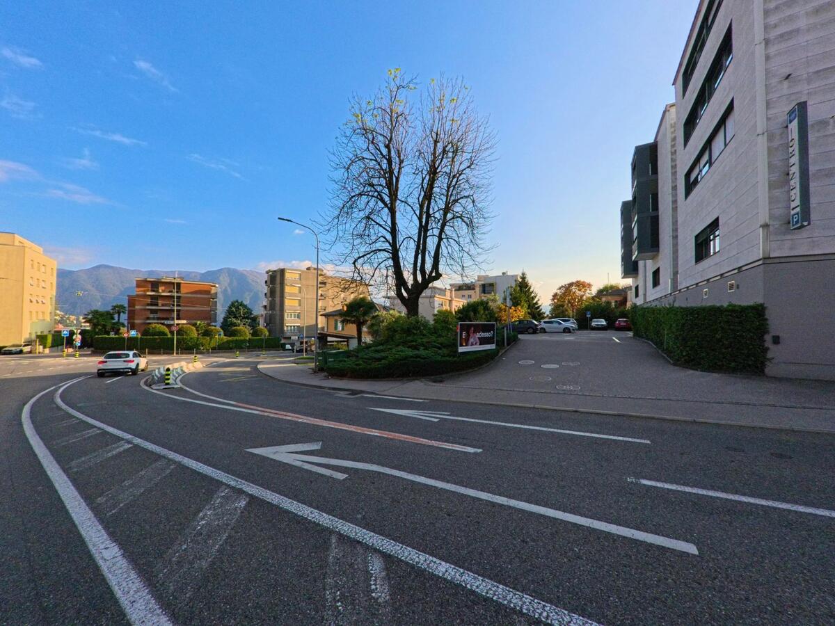 An einer sanft geschwungenen Strasse in Massagno (6900, Kanton Tessin) führt die Fahrbahn an Wohnbauten, einem einzelnen Baum und markierten Abbiegespuren vorbei.