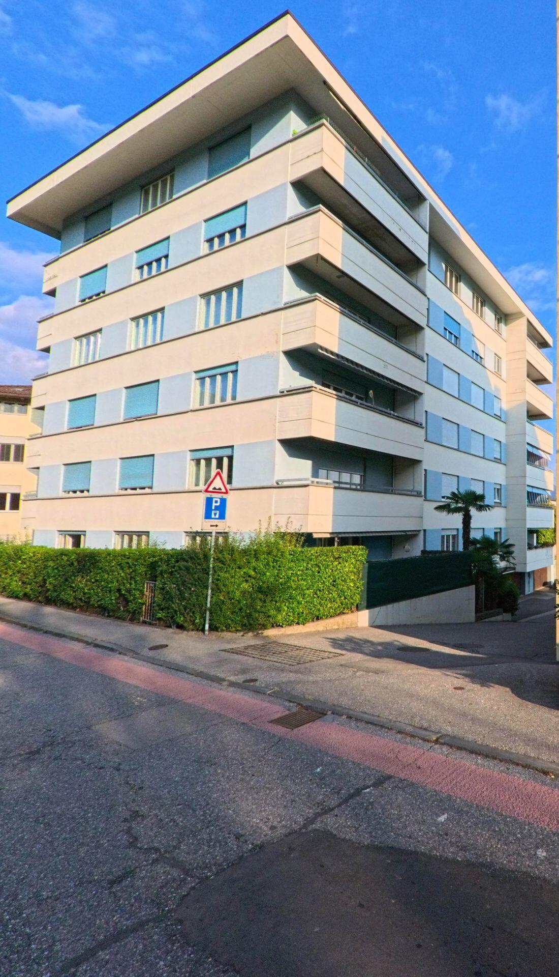 An einer leicht ansteigenden Strasse in Massagno (6900, Kanton Tessin) steht ein mehrgeschossiges Wohnhaus mit Balkonen, gepflegten Hecken und Zufahrt zur Tiefgarage.
