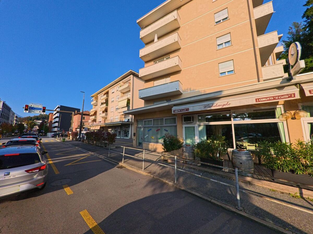 Entlang einer belebten Strasse in Massagno (6900, Kanton Tessin) reihen sich Wohnhäuser mit Balkonen an ein Geschäftslokal im Erdgeschoss bei sonnigem Tageslicht.