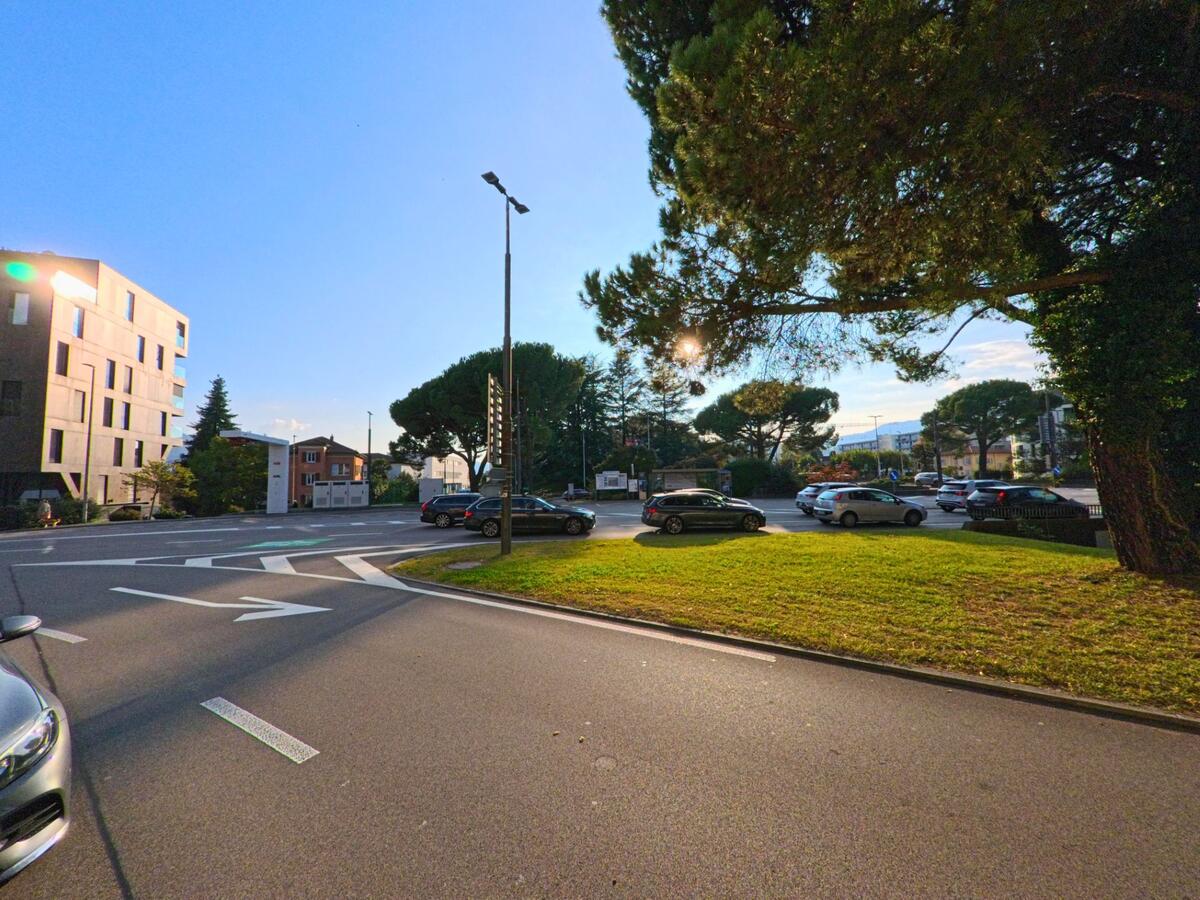 Neben einer begrünten Verkehrsinsel verläuft in Massagno (6900, Kanton Tessin) eine mehrspurige Strasse mit Abbiegespuren, parkierenden Autos und umliegenden Wohnbauten.