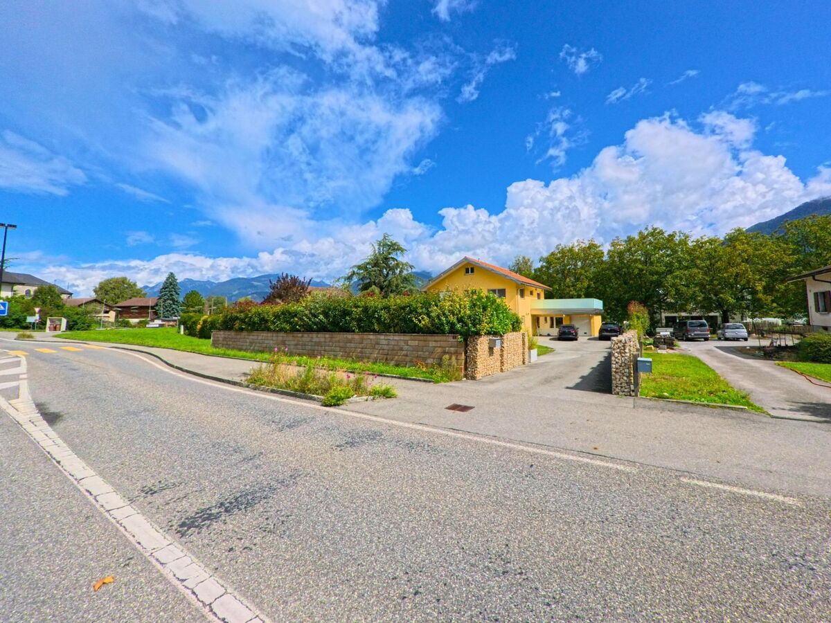 Ein gelbes Einfamilienhaus mit Einfahrt und Garten steht in Massongex (1869, Kanton Wallis) vor einer Kulisse von Bergen und Baeumen.
