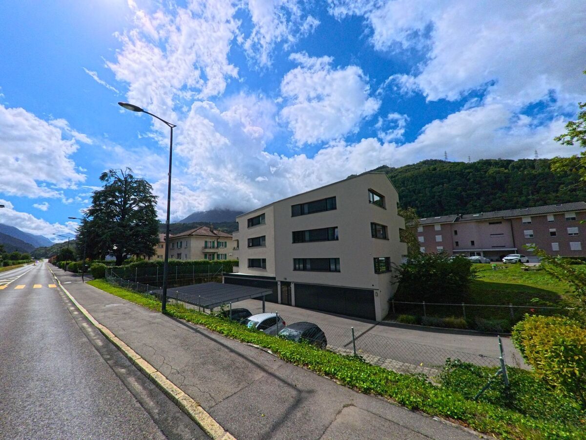 Ein modernes Wohnhaus mit Flachdach steht in Massongex (1869, Kanton Wallis) an einer langen Strasse mit Blick auf bewaldete Huegel.