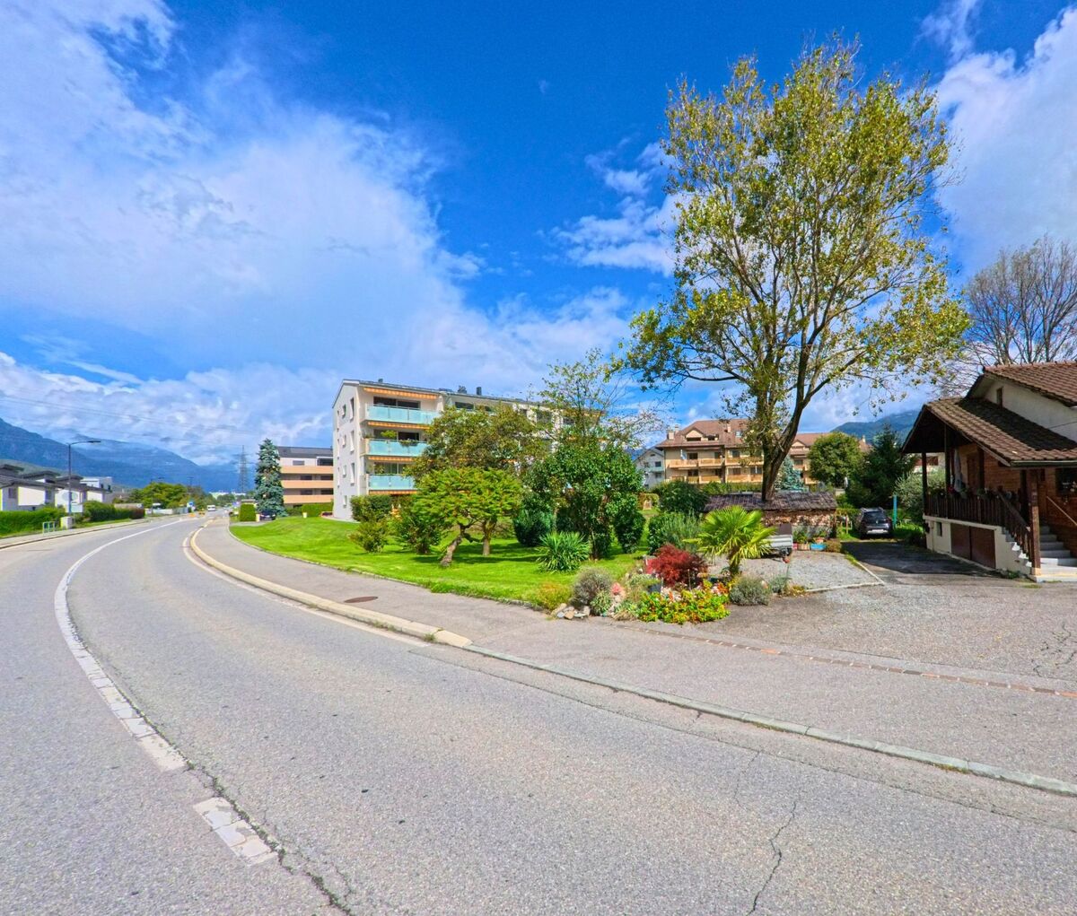 Eine geschwungene Strasse in Massongex (1869, Kanton Wallis) fuehrt an Mehrfamilienhaeusern und Gaerten mit grossen Baeumen vorbei.
