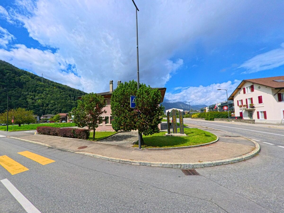 Eine kleine Verkehrsinsel mit Baeumen liegt in Massongex (1869, Kanton Wallis) an einer Hauptstrasse mit Wohnhaeusern.