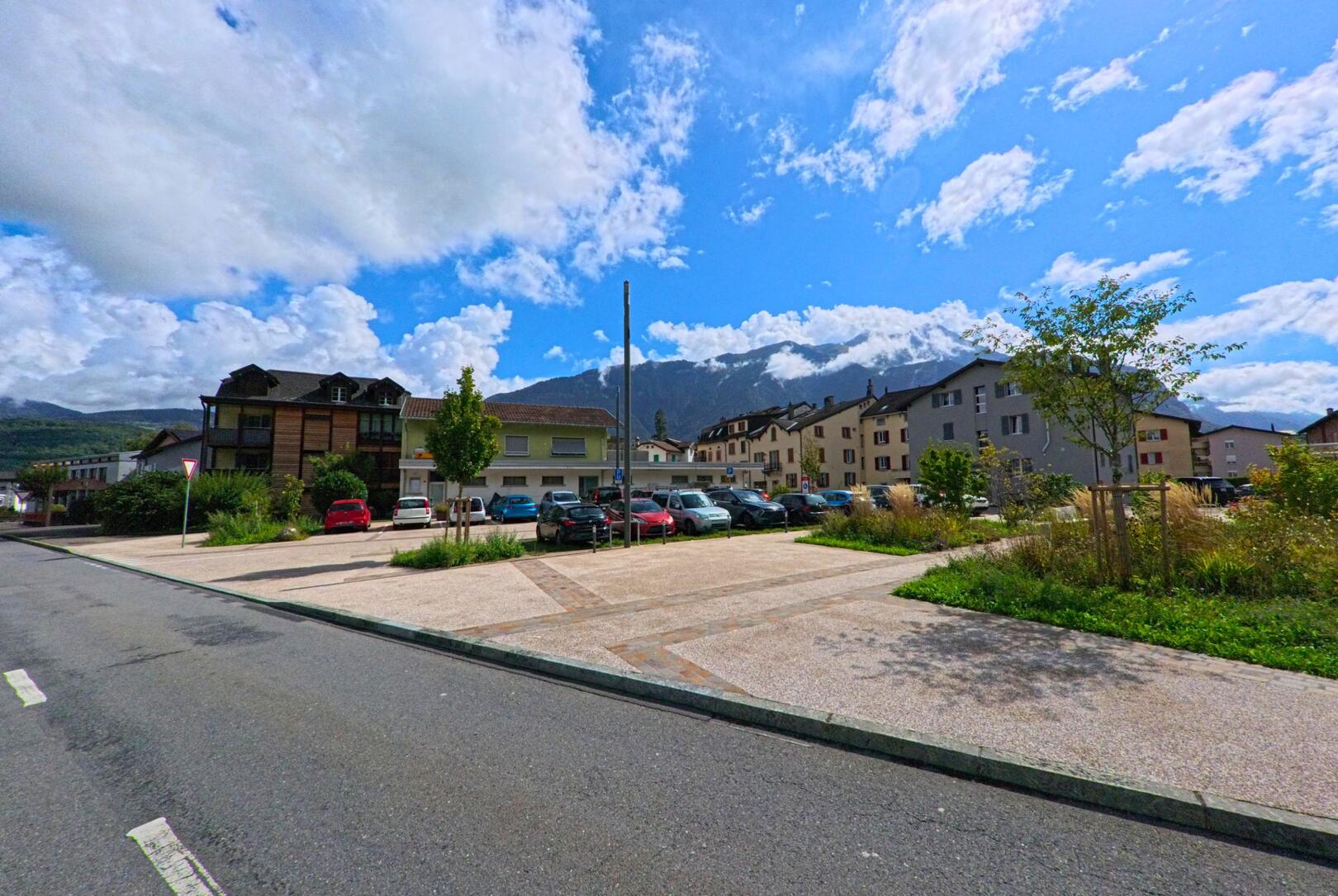 In Massongex (1869, Kanton Wallis) liegt ein zentraler Parkplatz von Wohnhaeusern umgeben, mit Bergen im Hintergrund.