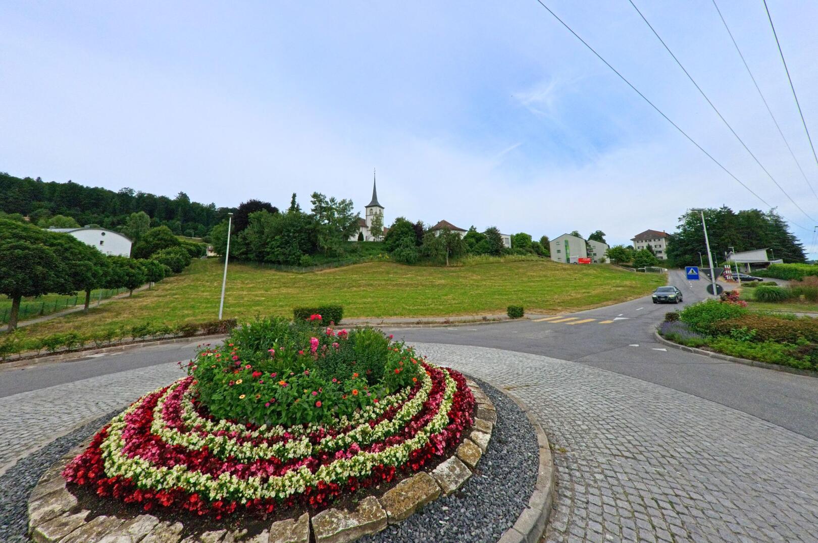 In Matran (1753, Kanton Freiburg) steht im Hintergrund eine Kirche auf einer Anhöhe, während im Vordergrund ein Kreisverkehr mit bunt bepflanztem Blumenbeet angelegt ist.