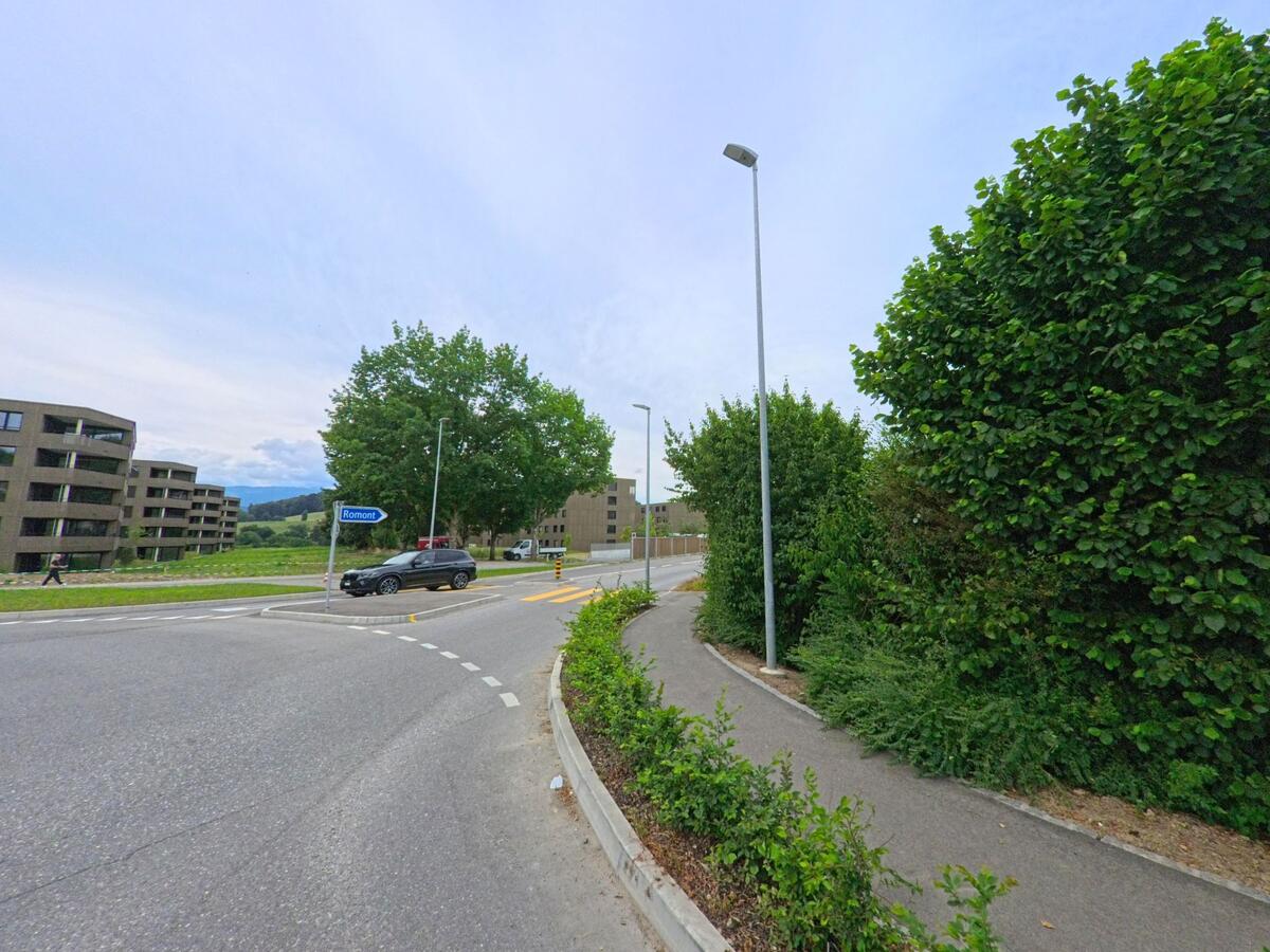 In Matran (1753, Kanton Freiburg) fuehrt eine Strasse mit Hinweisschild Richtung Romont vorbei an modernen Wohnbauten und einem baumbestandenen Fussweg.