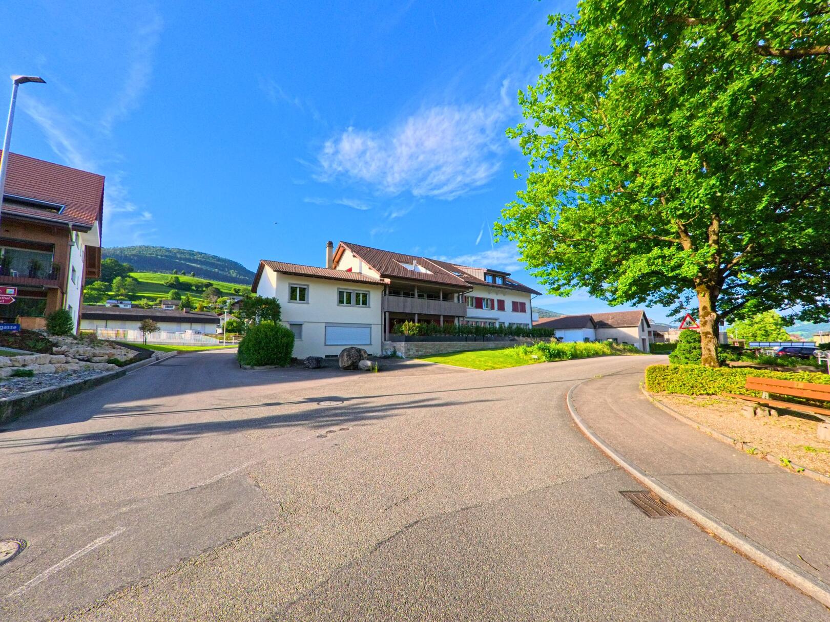 In Matzendorf (4713, Kanton Solothurn) verläuft eine Wohnstrasse an Einfamilienhäusern und einem grossen Baum mit Sitzbank vorbei.
