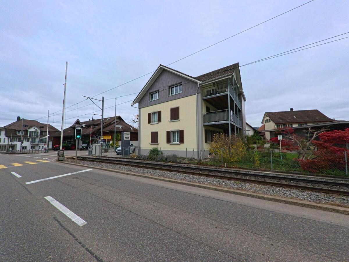 Neben den Bahngeleisen in Matzingen (9548, Kanton Thurgau) steht ein Wohnhaus mit Balkonen, während an der nahen Kreuzung eine Ampel auf Grün schaltet.