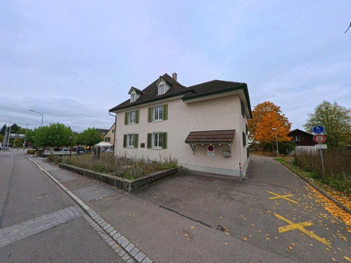 Am Rand eines Parkplatzes in Matzingen (9548, Kanton Thurgau) steht ein helles Wohnhaus mit grünen Fensterläden, daneben leuchtet ein Baum in herbstlichen Farben.