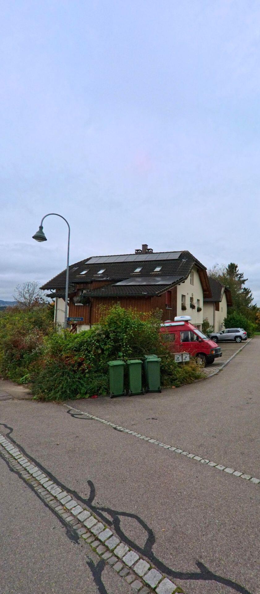 Am Rand einer Wohnstrasse in Matzingen (9548, Kanton Thurgau) steht ein Haus mit Solarpanels auf dem Dach, davor parkt ein rotes Fahrzeug neben grünen Abfallcontainern.