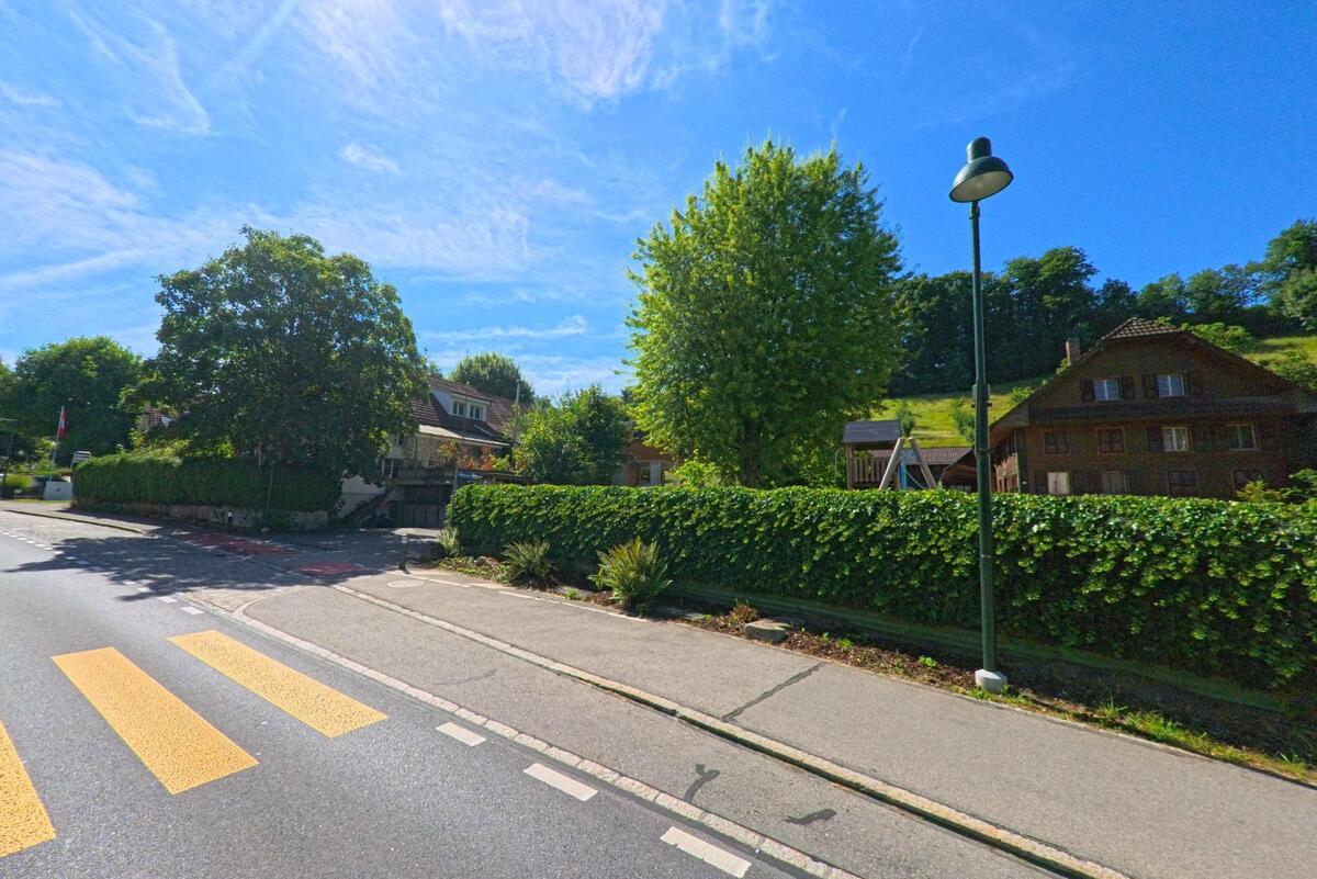 In Mauensee (6216, Kanton Luzern) fuehrt ein Fussgaengerstreifen ueber eine Hauptstrasse mit angrenzender Hecke, hinter der sich ein traditionelles Holzhaus und ein Spielplatz befinden.