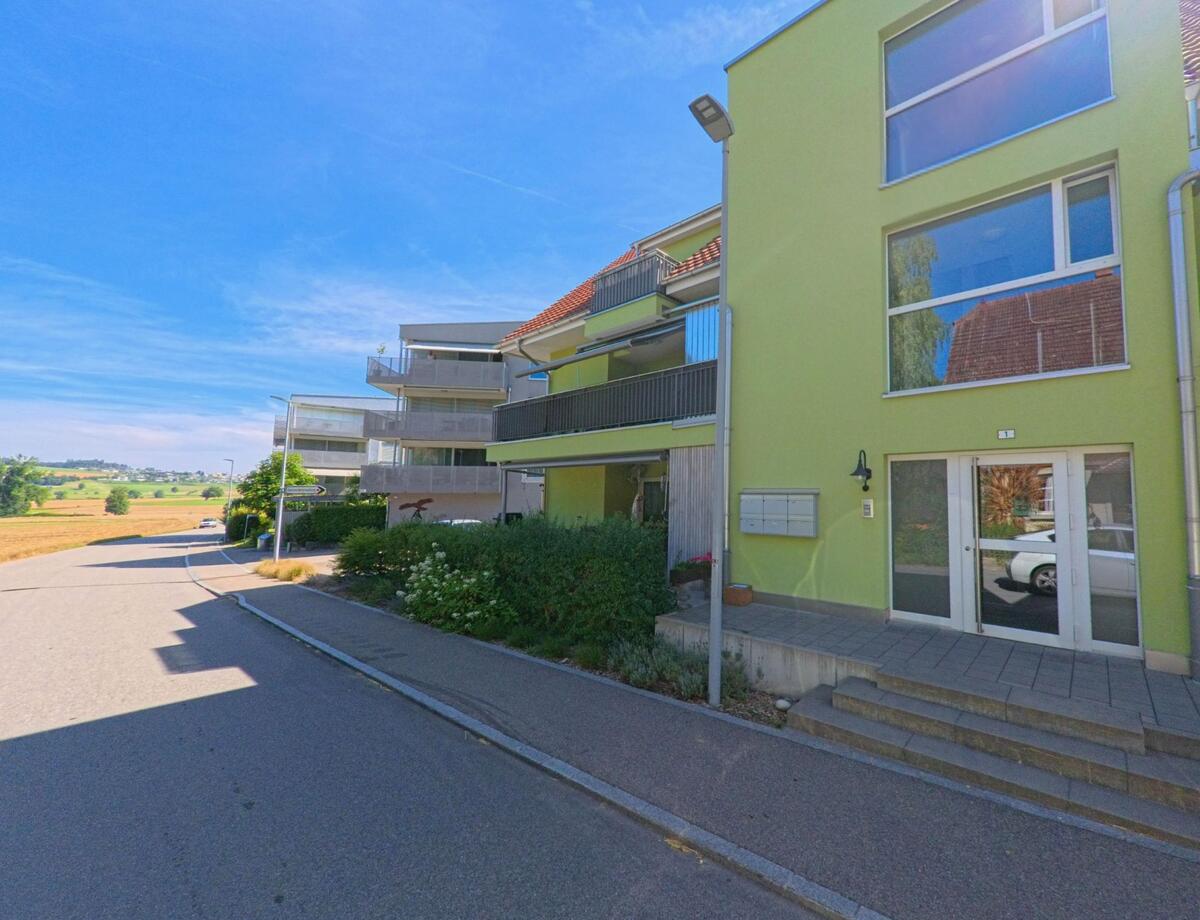 In Mauensee (6216, Kanton Luzern) steht ein modernes Wohnhaus mit gruener Fassade und Balkonen an einer Quartierstrasse mit Blick auf offene Felder.