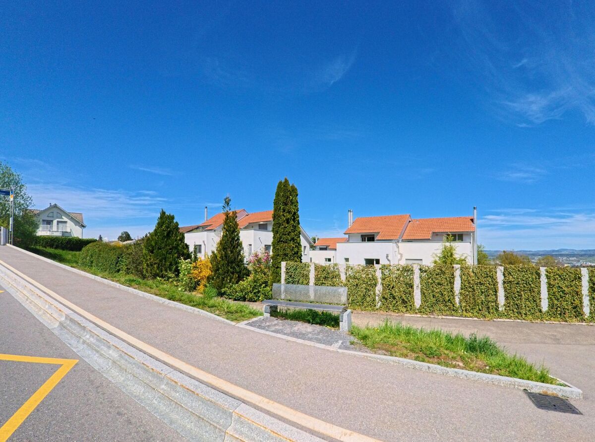Von einer leicht ansteigenden Strasse in Maur (8122, Kanton Zürich) bietet sich ein Blick auf gepflegte Wohnhäuser mit roten Dächern hinter einer Hecke und auf das hügelige Umland.