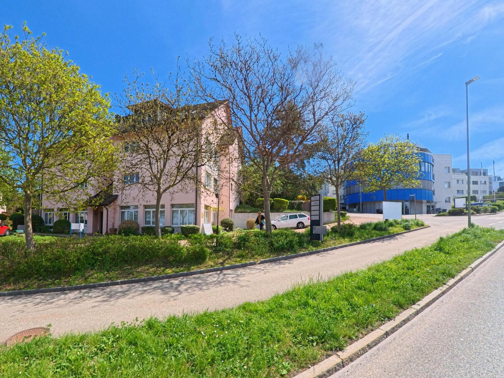 An einer leicht ansteigenden Strasse in Maur (8122, Kanton Zürich) stehen mehrstöckige Wohn- und Bürogebäude, gesäumt von jungen Bäumen und Grünstreifen.