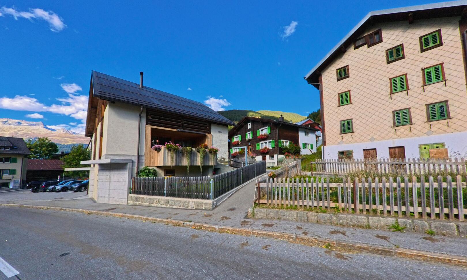In Medel (Lucmagn, 7184, Kanton Graubuenden) stehen traditionelle Holzhäuser mit grünen Fensterläden entlang einer ruhigen Strasse, umgeben von den Bergen im Hintergrund.