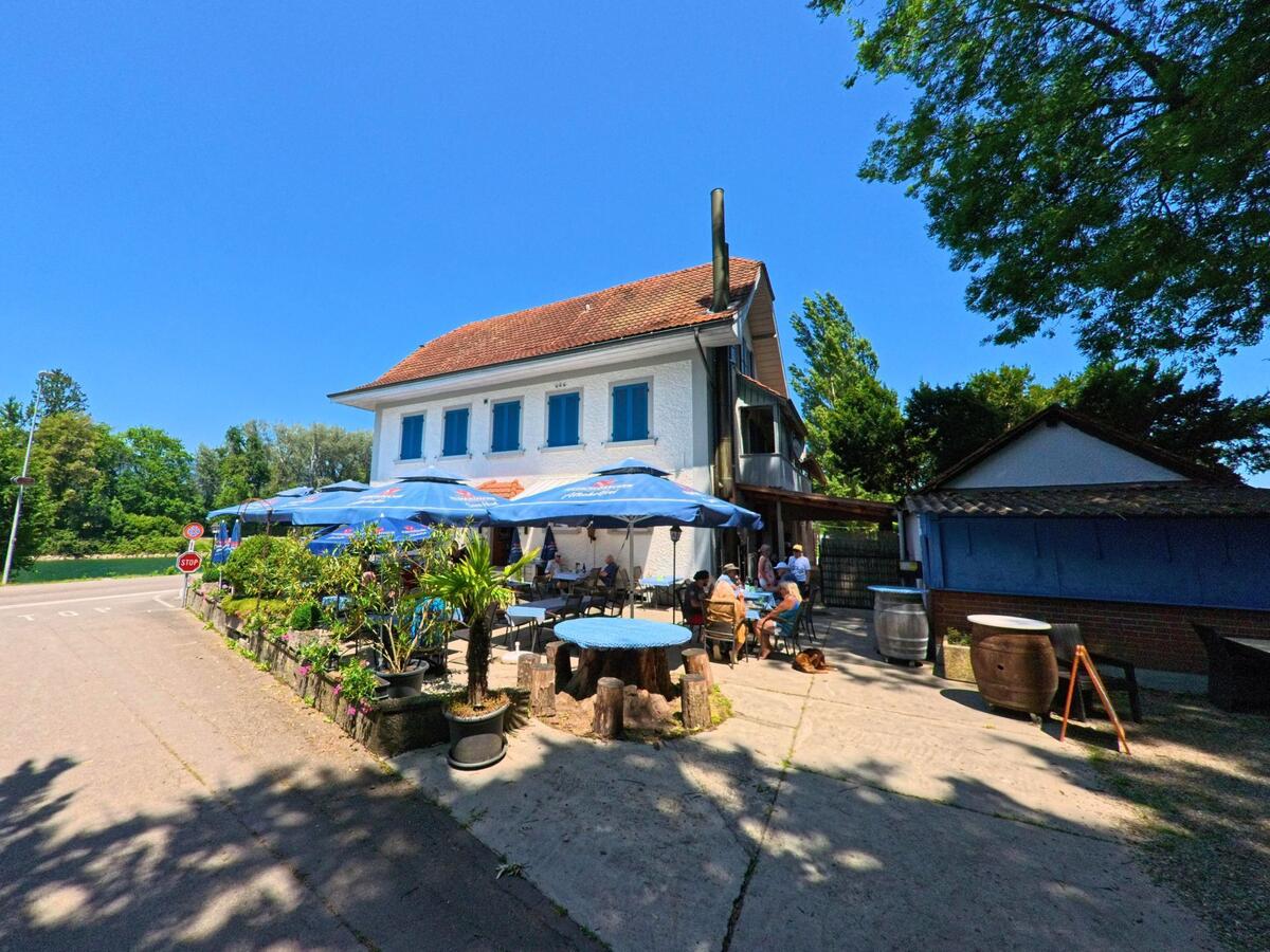 In Meienried (2553, Kanton Bern) geniessen Gäste bei Sonnenschein ihren Aufenthalt unter blauen Sonnenschirmen vor einem weissen Gebäude mit roten Ziegeln.
