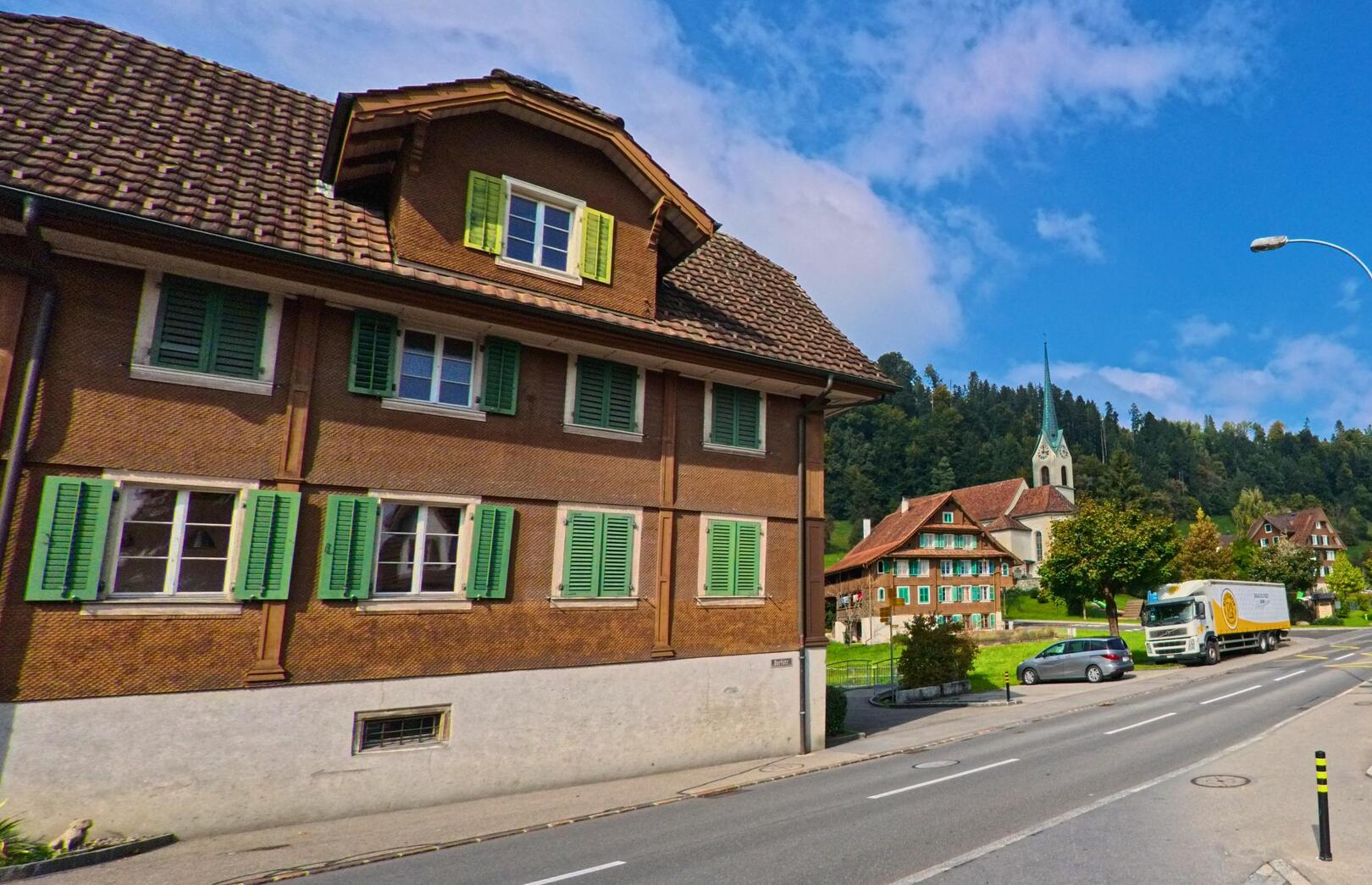 In Meierskappel (6344, Kanton Luzern) steht ein traditionelles Gebäude mit grünen Fensterläden entlang der Straße, während im Hintergrund die Kirche und ein weiteres historisches Gebäude zu sehen sind.