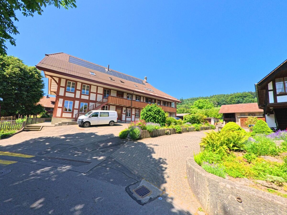 In Meikirch (3045, Kanton Bern) ist ein traditionelles Fachwerkhaus zu sehen, umgeben von einem gepflegten Innenhof mit Blumenbeeten und einem Lieferwagen im Vordergrund.