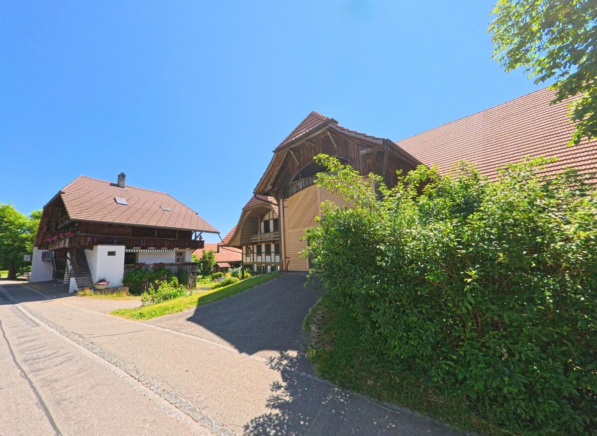 In Meikirch (3045, Kanton Bern) ist eine Straße zu sehen, die an einem traditionellen Fachwerkhaus mit Blumen auf dem Balkon und einem modernen Gebäude mit rotem Ziegeldach vorbeiführt.