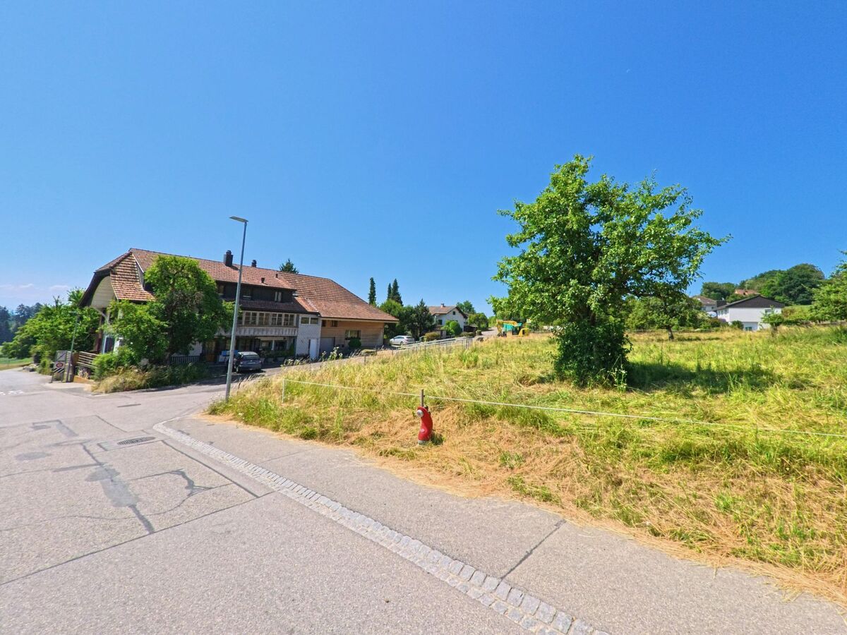 In Meikirch (3045, Kanton Bern) führt die Straße an einem historischen Gebäude mit einem gut gepflegten Garten vorbei, in der Nähe eines offenen Feldes.