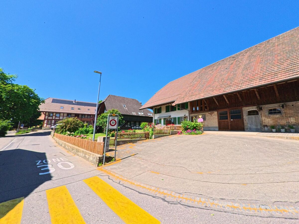 In Meikirch (3045, Kanton Bern) ist eine Straße mit einem historischen Gebäude und einem großen, roten Ziegeldach abgebildet, daneben ein Verkehrsschild für die 30 km/h Zone.