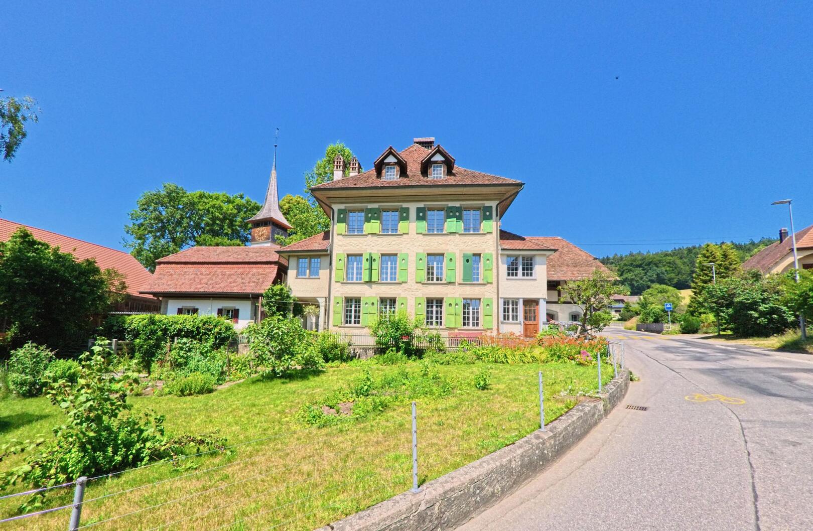 In Meikirch (3045, Kanton Bern) führt die Strasse an einem stattlichen Gebäude mit grünen Fensterläden und einem gepflegten Garten vorbei, umgeben von Bäumen und Wiesen.