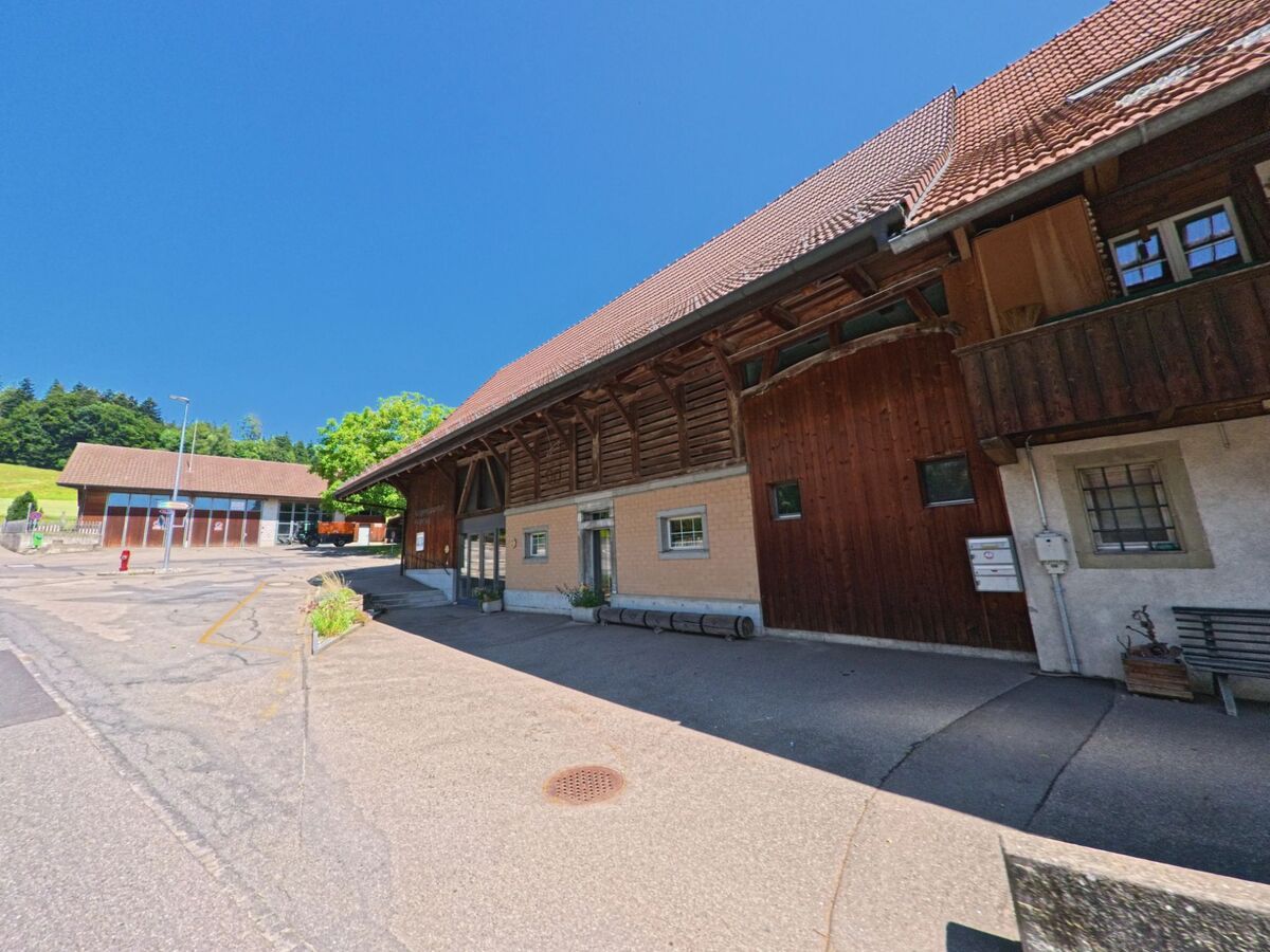 In Meikirch (3045, Kanton Bern) sind zwei Gebäude zu sehen, eines mit Holzfassade und das andere mit einem modernen Bau, in der Nähe eines ruhigen Straßeneckenbereichs.
