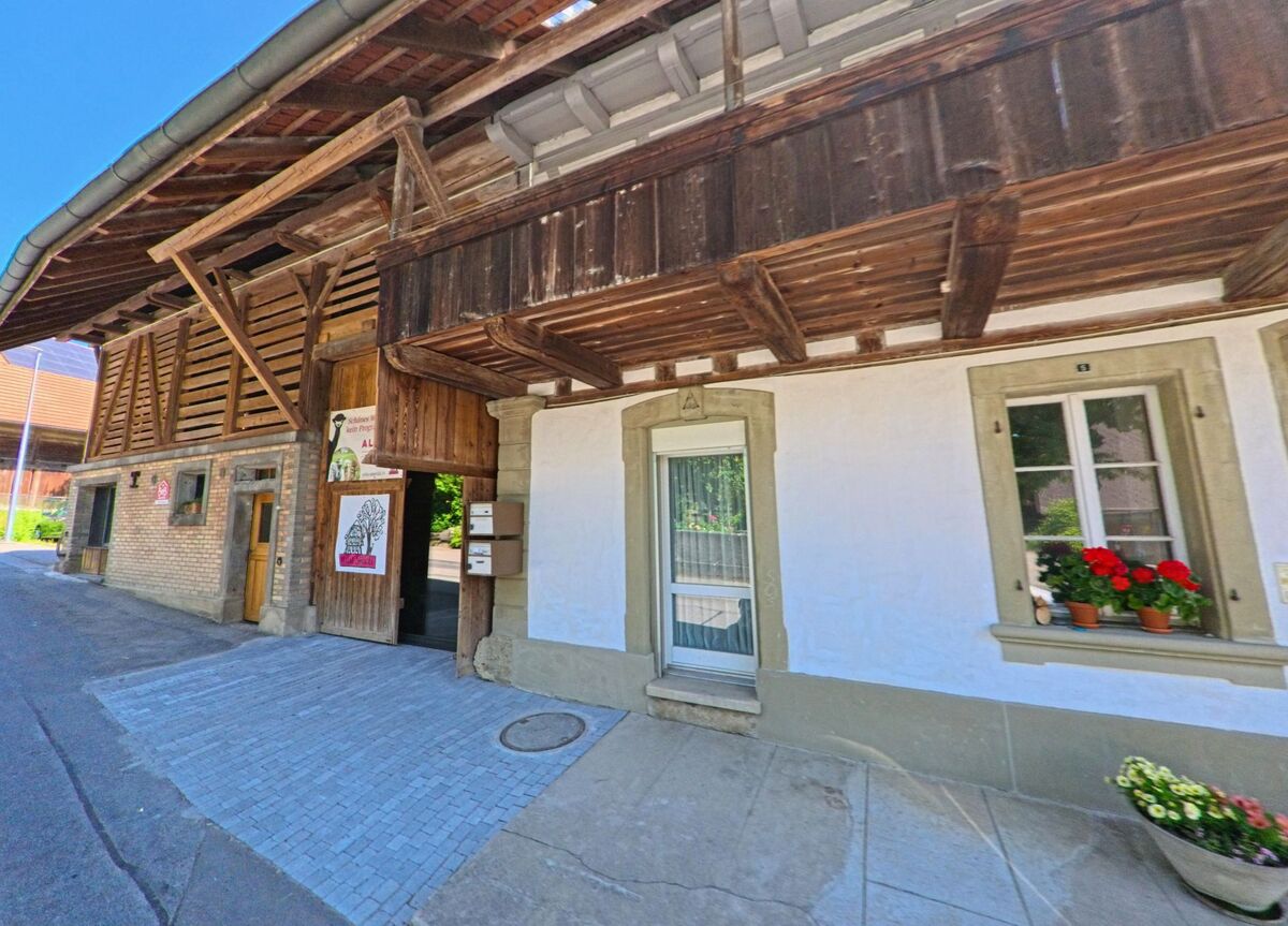 In Meikirch (3045, Kanton Bern) ist ein historisches Gebäude mit Holzverkleidung und Blumen am Fenster zu sehen, das in eine ruhige Straßenszene eingebettet ist.