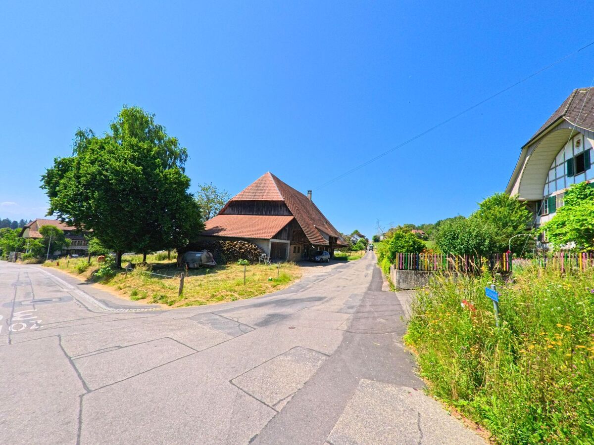 In Meikirch (3045, Kanton Bern) zeigt die Straße eine Abzweigung, die von einem historischen Gebäude mit einem großen Dach und grünen Wiesen flankiert wird.