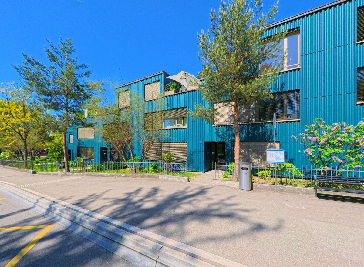 Ein modernes Gebäude mit blauer Fassadenverkleidung steht in Meilen, PLZ 8706 im Kanton Zürich neben einer ruhigen Strasse mit Bäumen und Sitzbank.