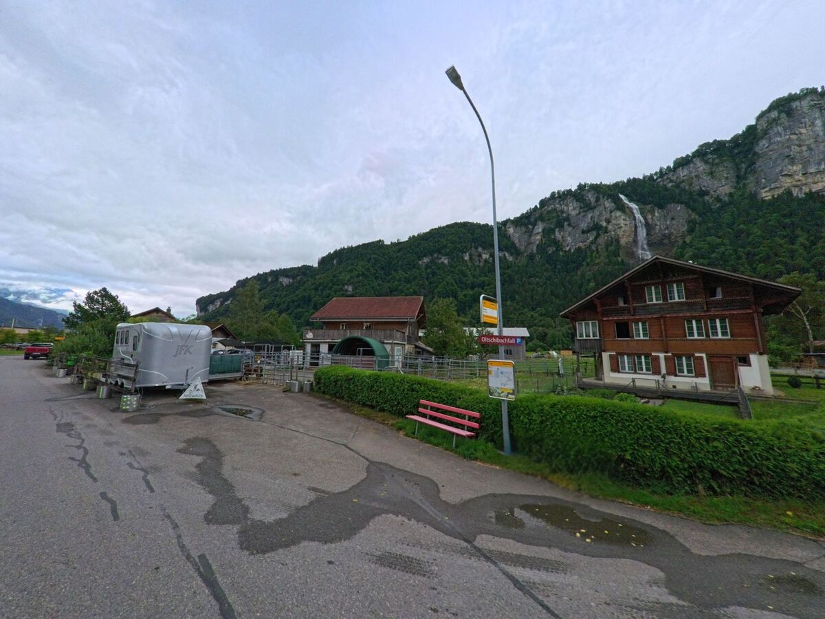 In Meiringen (3857, Kanton Bern) liegt eine Posthaltestelle vor einem traditionellen Bauernhof, während im Hintergrund ein Wasserfall steil aus der Felswand herabstürzt.