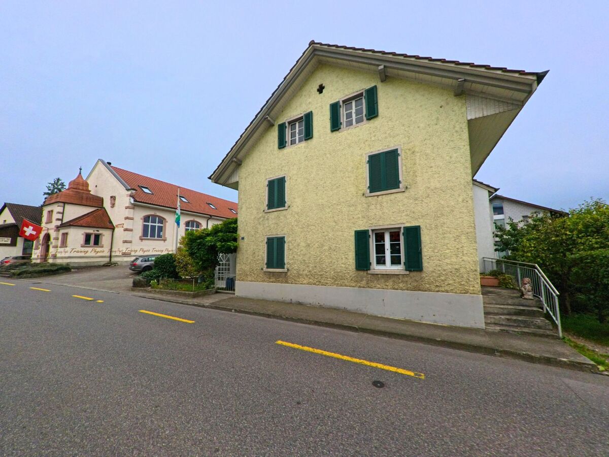 Ein helles Wohnhaus mit grünen Fensterläden steht an der Hauptstrasse in Meisterschwanden (5616, Kanton Aargau), daneben ein Gebäude mit rotem Dach und Schweizer Fahne.