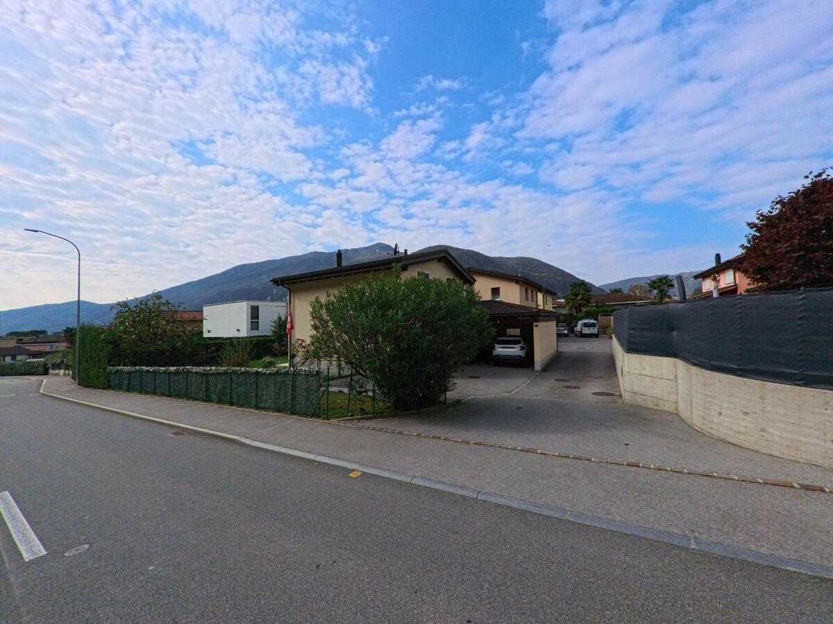 Ein Einfamilienhaus mit Einfahrt und Hecken liegt an einer ruhigen Strasse in Melano (6818, Kanton Tessin), während sich dahinter bewaldete Hügel erheben.