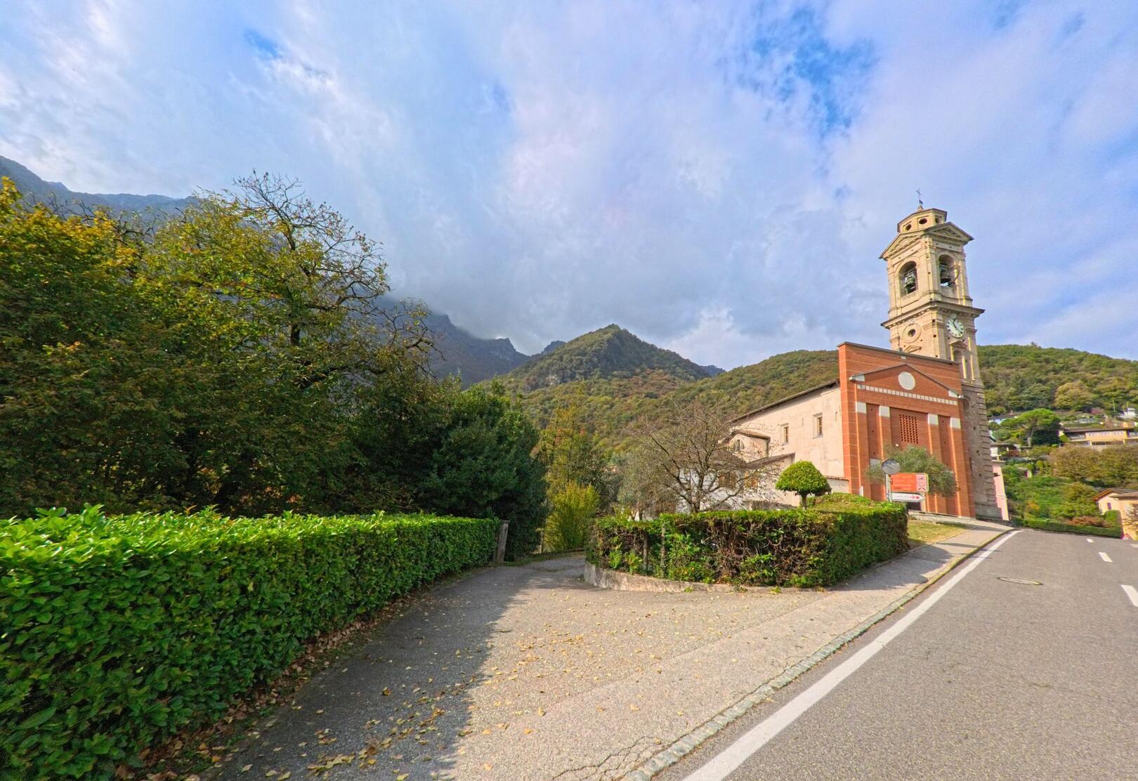Am Rand einer Strasse mit Hecken und Gehweg steht die Kirche mit Glockenturm in Melano (6818, Kanton Tessin) vor bewaldeten Hügeln unter hellem Himmel.