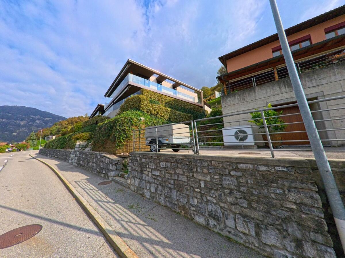 Moderne Wohnhäuser mit Glasbalkonen stehen oberhalb einer Steinmauer entlang der Strasse in Melano (6818, Kanton Tessin) vor einem bewaldeten Hang.